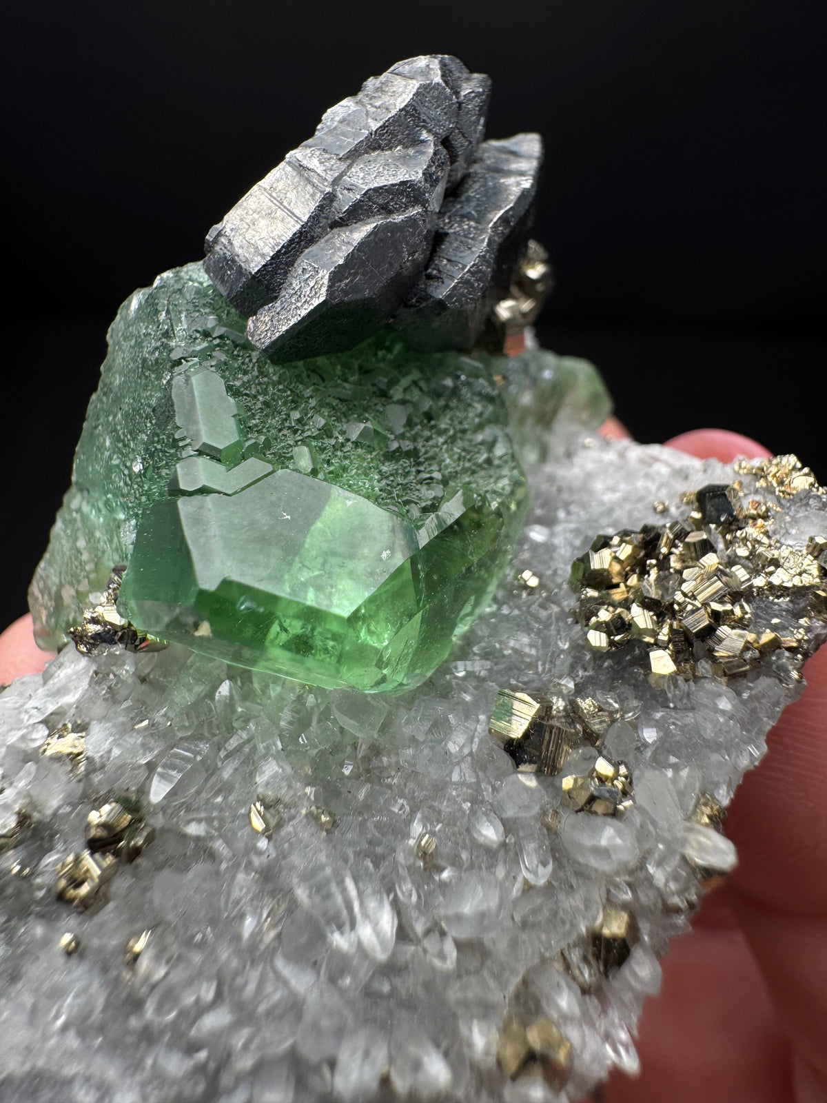 A hand holding a mineral specimen featuring a prominent green faceted crystal, a cluster of dark metallic gray crystals, and a clear quartz-like matrix with scattered gold-colored crystals, set against a solid black background.